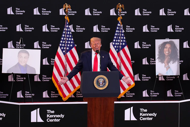 A man in a suit standing at a lectern in front of two US flags with enlarged photos on either side.