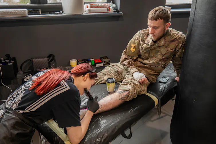 A person with long, red hair tattoos a man's shin