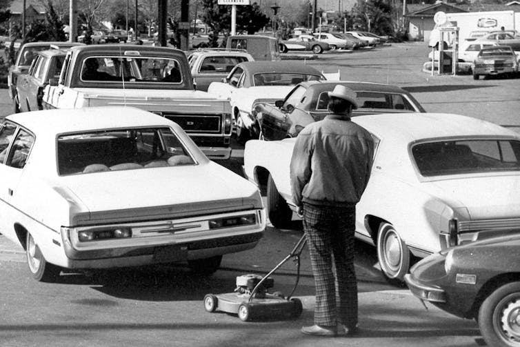 a black-and-white photo shows classic cars and a man pushing a lawnmower in a long line on the road