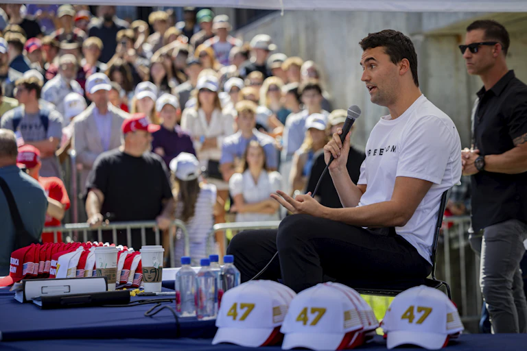 A white man with short brown hair wearing a white shirt, speaking into a microphone before a crowd of people