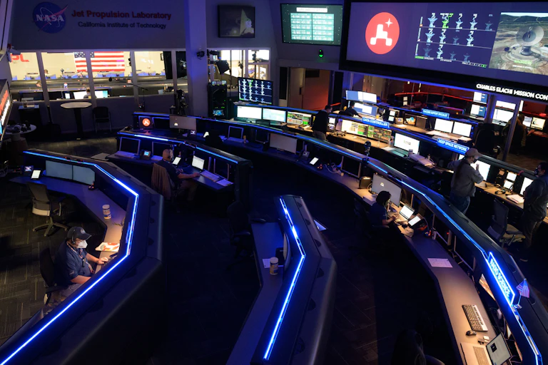 A group of scientists sitting in a dark control room, looking at large screens at the front of the room