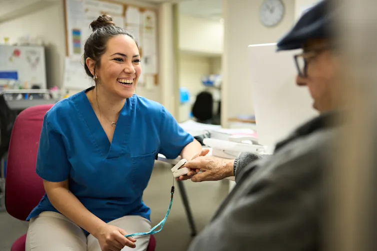 A nurse sits with an older man, holds his hand while measuring his blood oxygen levels.