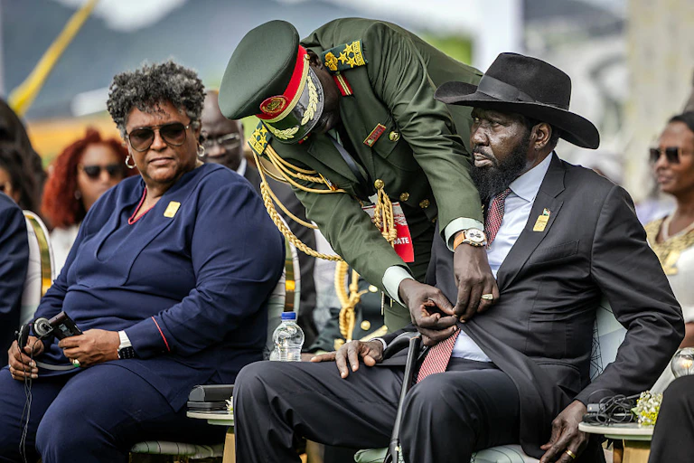 A seated man with his hands out as another in army fatigues leans over him to unbutton his jacket