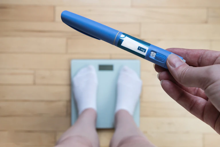 A person standing on a scale holds a blue vial of semaglutide.