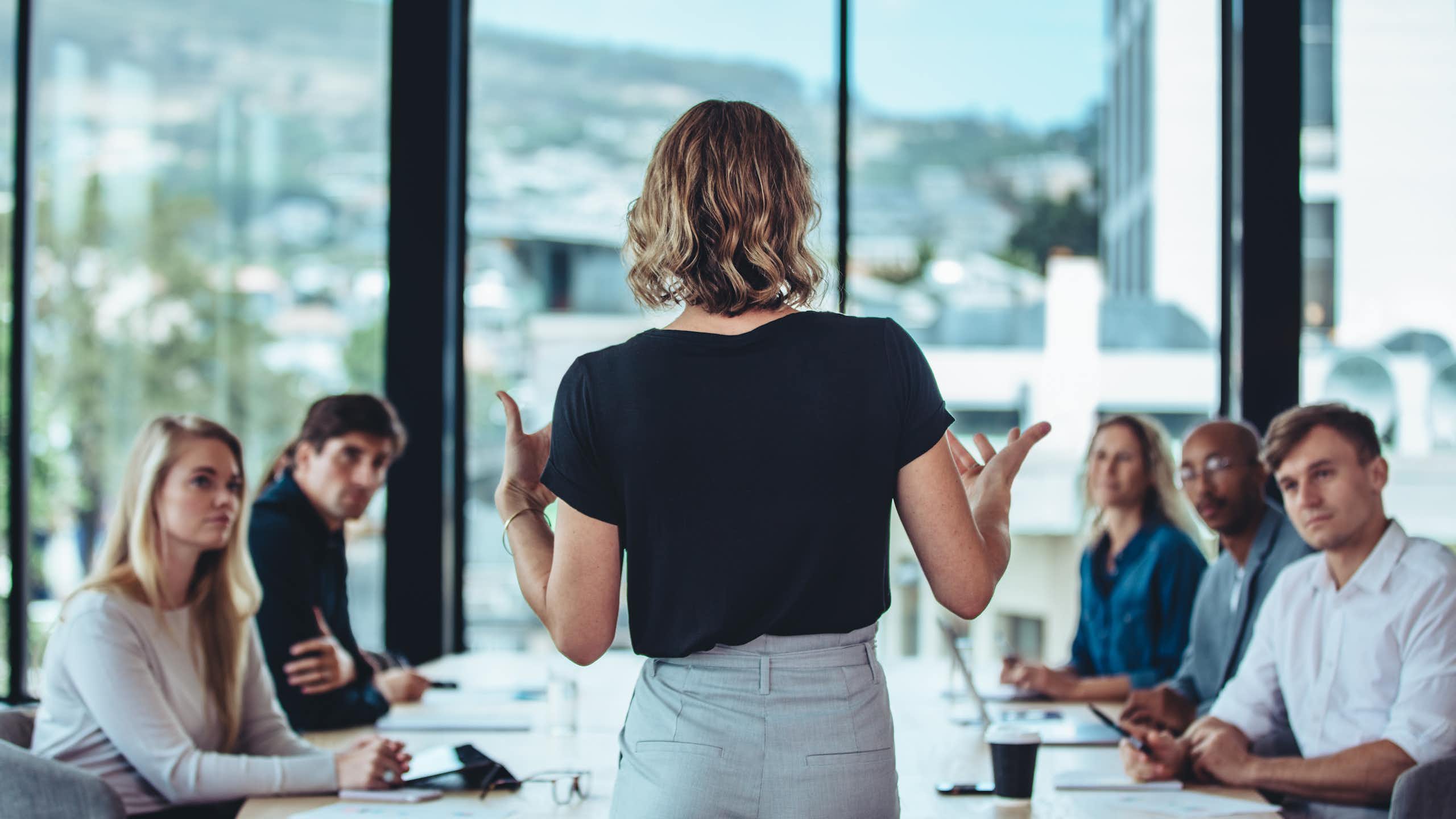 A woman wearing a black shirt is standing and explaining something in front of a rectangular table where five or six people are sitting and looking at her.