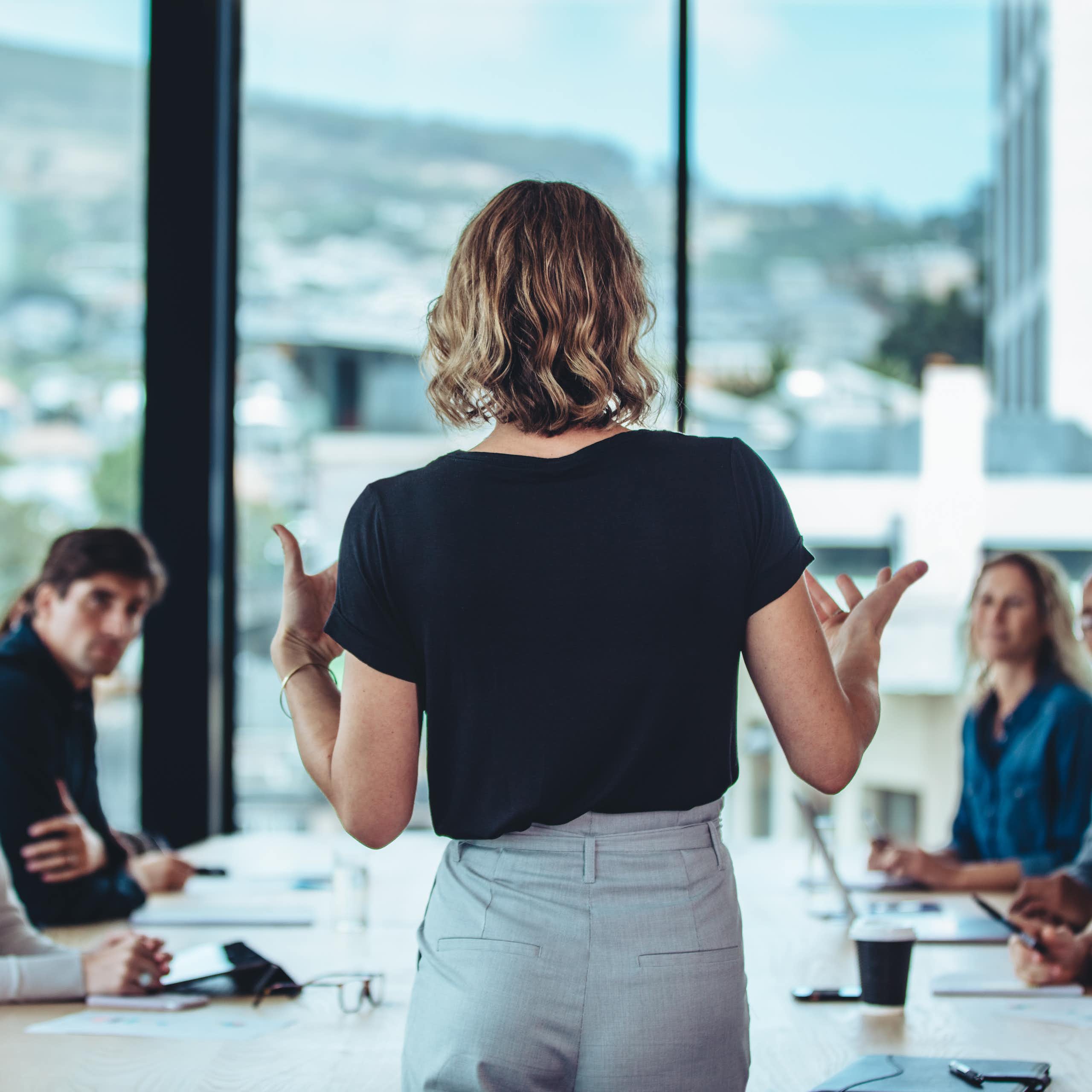 A woman wearing a black shirt is standing and explaining something in front of a rectangular table where five or six people are sitting and looking at her.