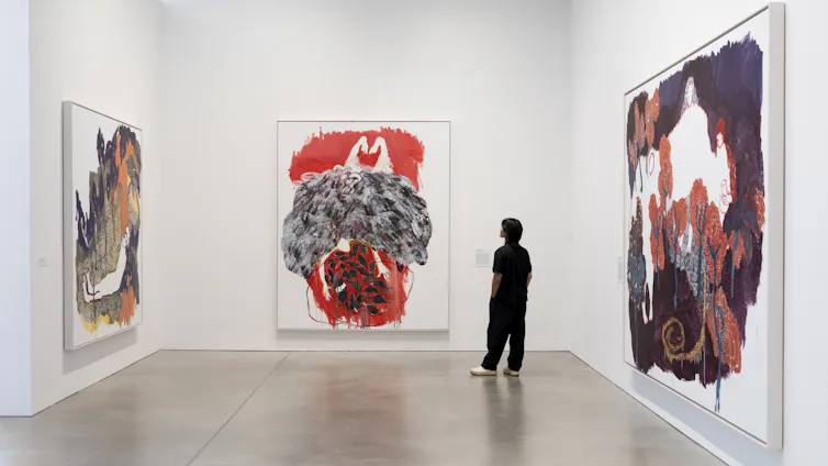 An art gallery with white walls and three vast red, black and white paintings hanging, looked at by a person dressed in black.