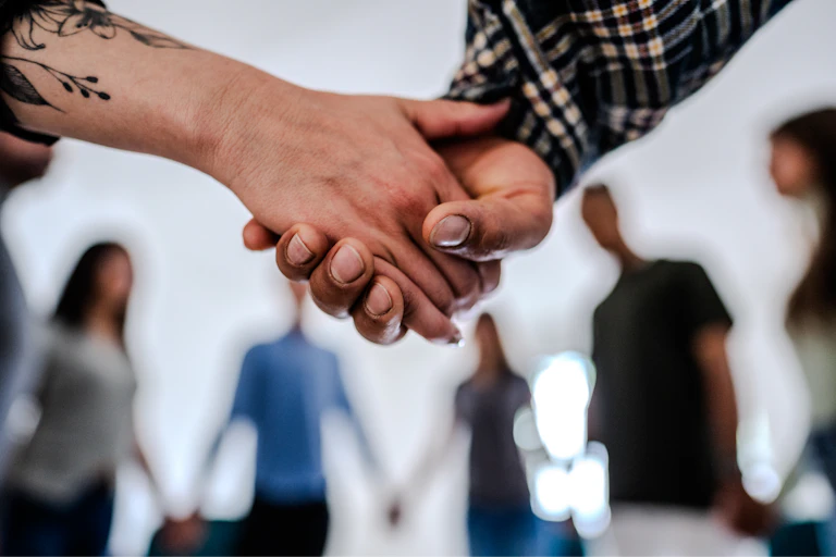 Two people's hands clasped together in foreground with blurred people in background seen standing in circle and holding hands