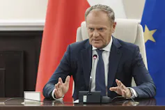 A balding man in a suit sits at a desk gesturing as he speaks.