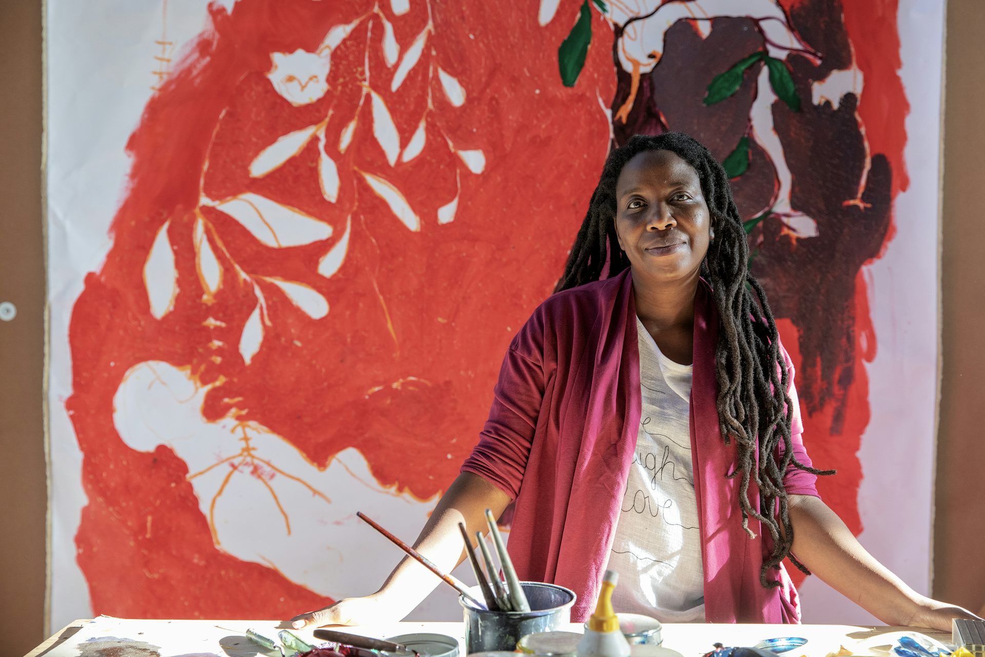 An African woman with long dreadlocks smiles as she looks into camera, standing in front of a table with paints and brushes, behind her a large red painting.