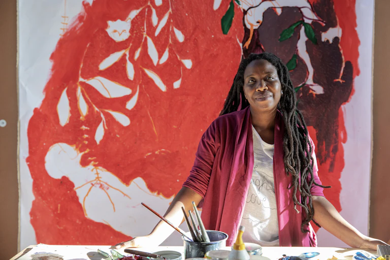 An African woman with long dreadlocks smiles as she looks into camera, standing in front of a table with paints and brushes, behind her a large red painting.