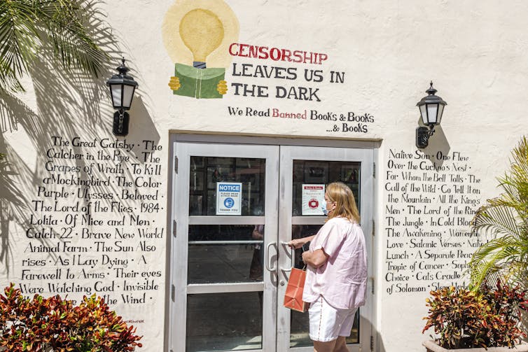 A woman wears a light-colored shirt and holds a door handle of a building that has painted words on its light wall: 'Censorship leaves us in the dark.'
