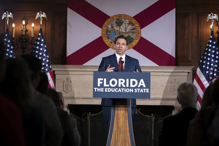 A white man with dark hair wears a navy blue suit and stands at a podium that says 'Florida, the education state.'