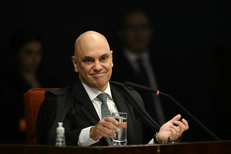 Bald man in a judge's black robe, a white dress shirt and a blue tie, smirks for the camera.