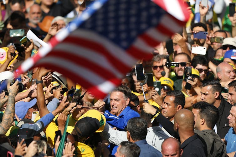 A man in a blue jacket is thronged by a crowd dressed in yellow and green below a waving American flag.