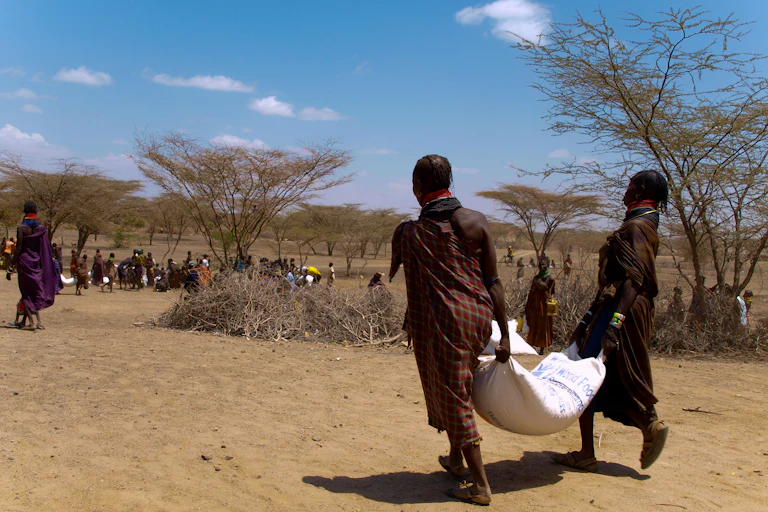 Two women carrying a sack of food between them while walking towards a larger group of women doing the same thing in a dry, arid area