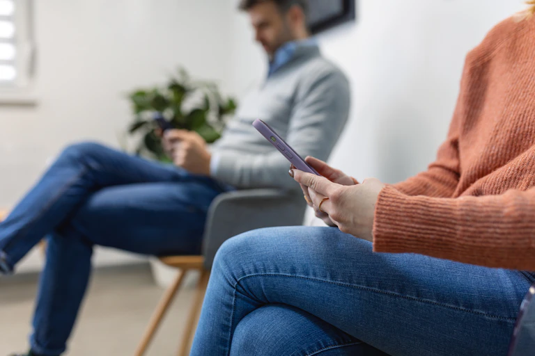 Woman and man sitting on chairrs in waiting room, scrolling on phone