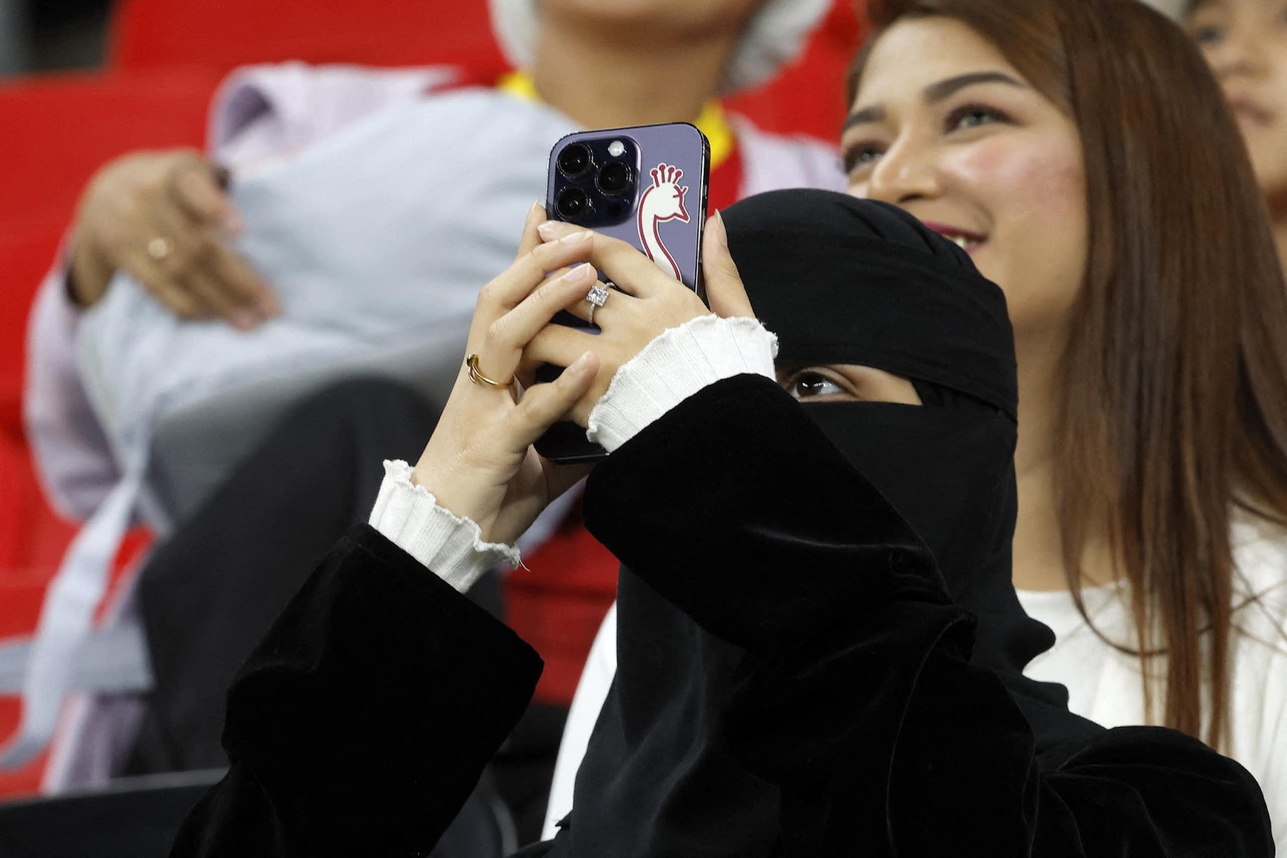 A woman wearing a niqab holds up a smartphone with a sticker of a bird on it. A woman with uncovered hair is visible behind her.