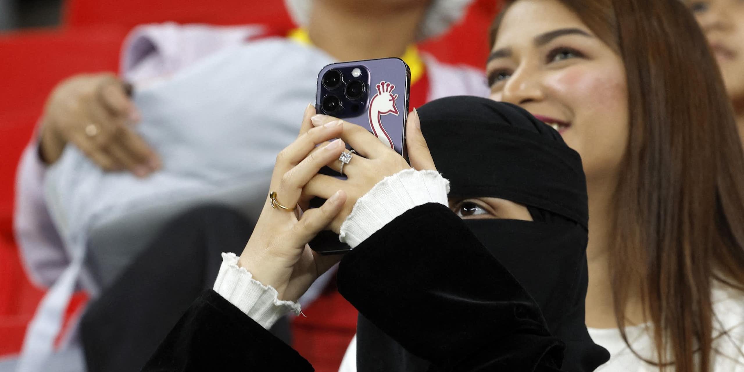 A woman wearing a niqab holds up a smartphone with a sticker of a bird on it. A woman with uncovered hair is visible behind her.