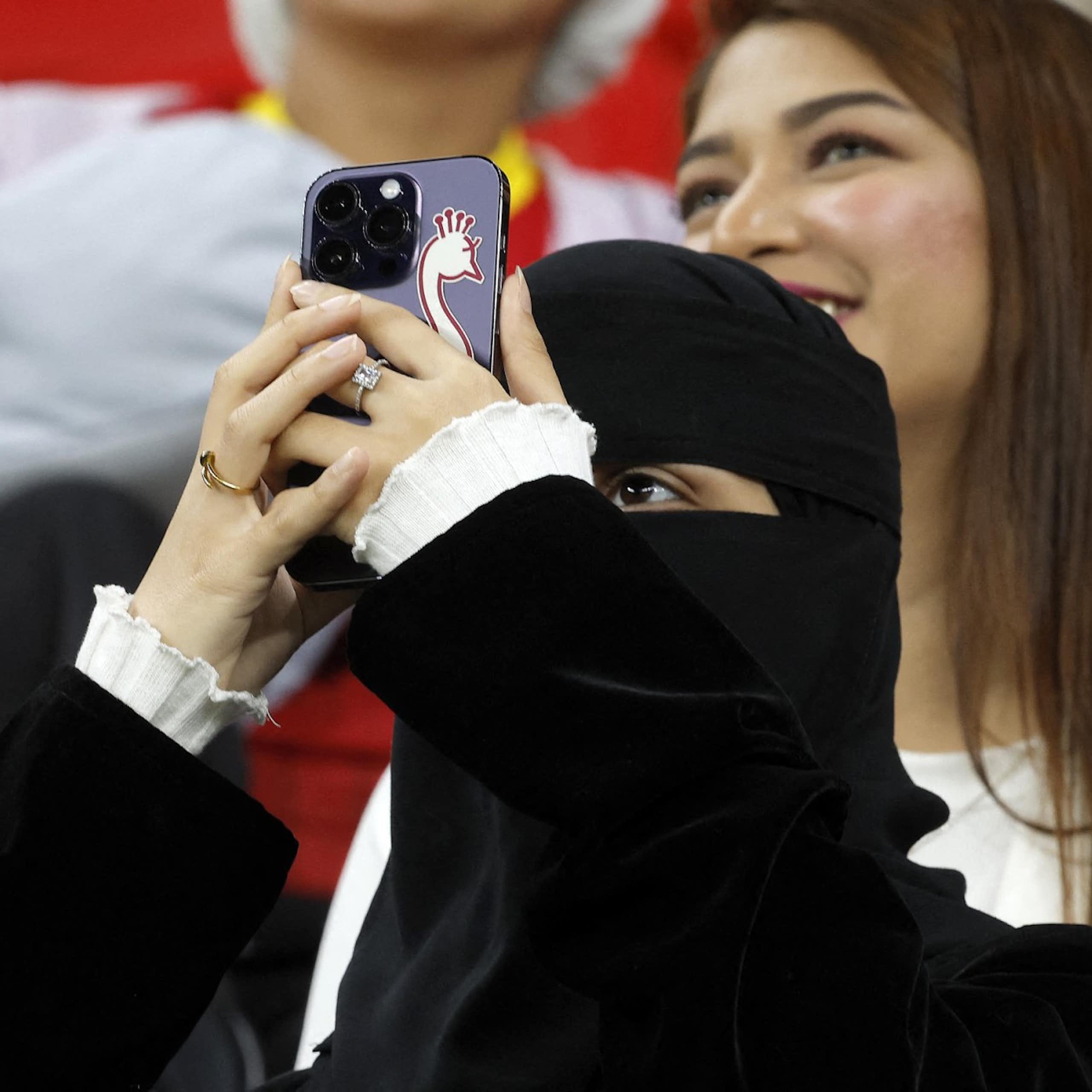 A woman wearing a niqab holds up a smartphone with a sticker of a bird on it. A woman with uncovered hair is visible behind her.