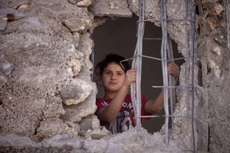 A young Palestinian girl looks out through a hole in the wall of her family home.