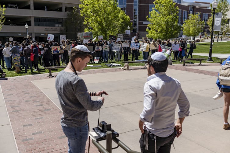 Two men wearing kippahs stand together in front of a crowd of people who are holding signs.