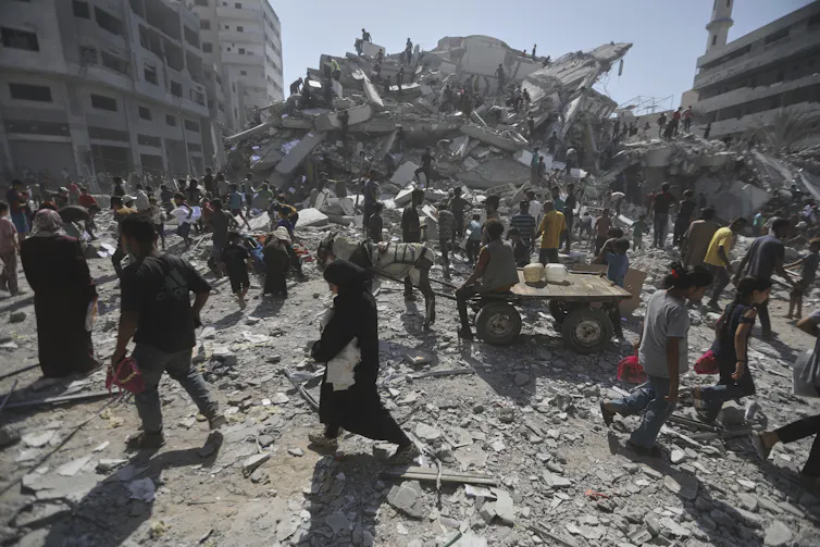 People look at the rubble of a building with mountains of rubble around them.