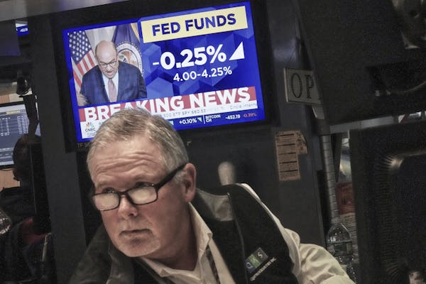 a bespectacled white man in a vest look on as a tv screen shows news of fed rate cut behind him