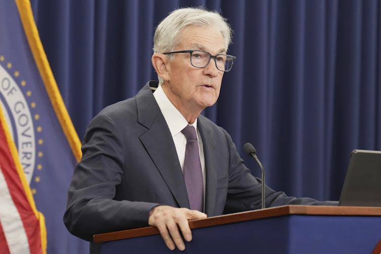 a bespectacled white man in a suit stands before a podium with a micrphone