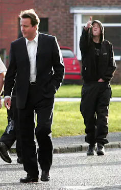 A teenager makes a gun sign with his hands behind David Cameron's back.