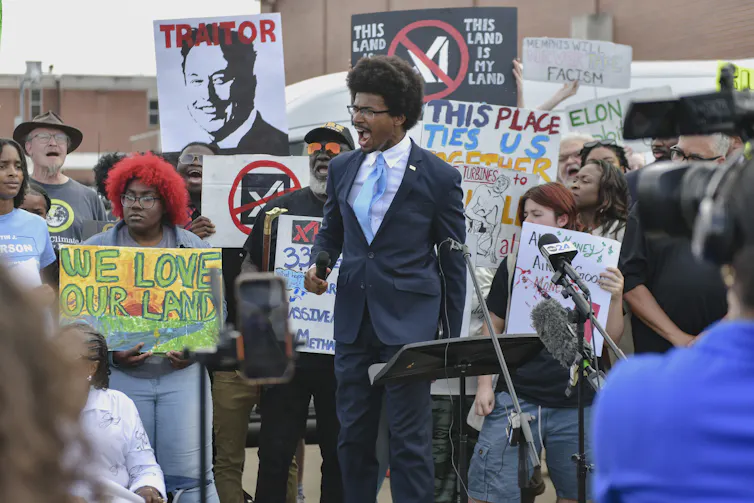 A man in a suit and tie stands in front of a crowd, speaking while they hold signs.
