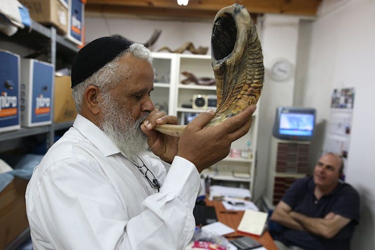 A man in a black skullcap and white shirt, who has a long white beard, blows into a large animal horn.