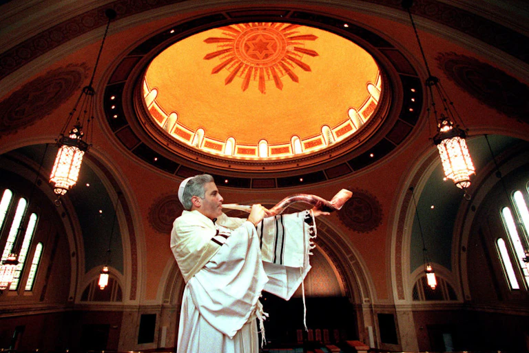 A man in white robes blows into a long animal horn as he stands in the center of an ornate sanctuary