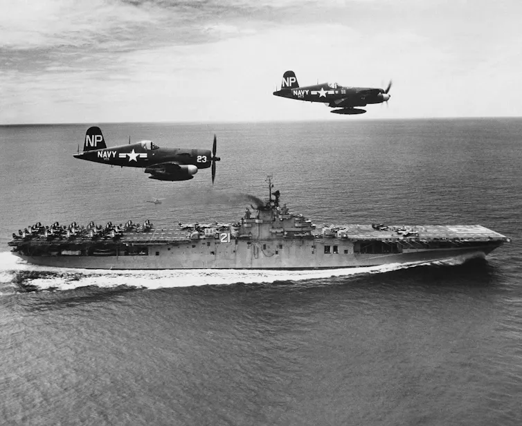 A black-and-white photo shows two aircraft flying over an aircraft carrier.