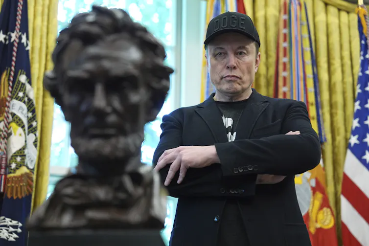 Elon Musk portant une casquette se tient dans le Bureau ovale