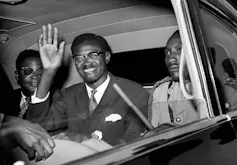A Black man smiles and waves from the back of a car.