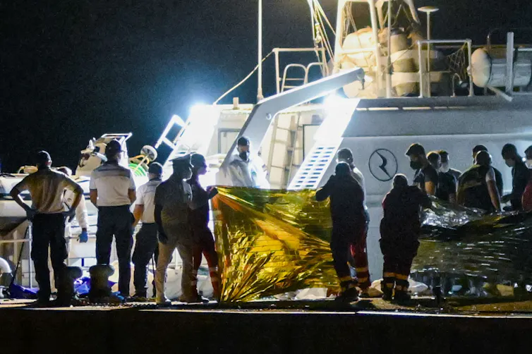 A dock at night with rescue workers holding up a yellow tarp to shield bodies.