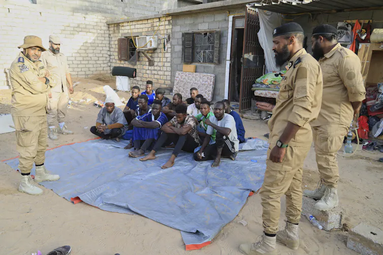 A group of Black people sit on a blue tarp surrounded by armed police officers.