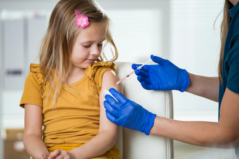 Nurse wearing surgical gloves gives a shot to a school-age girl who sits in a chair.