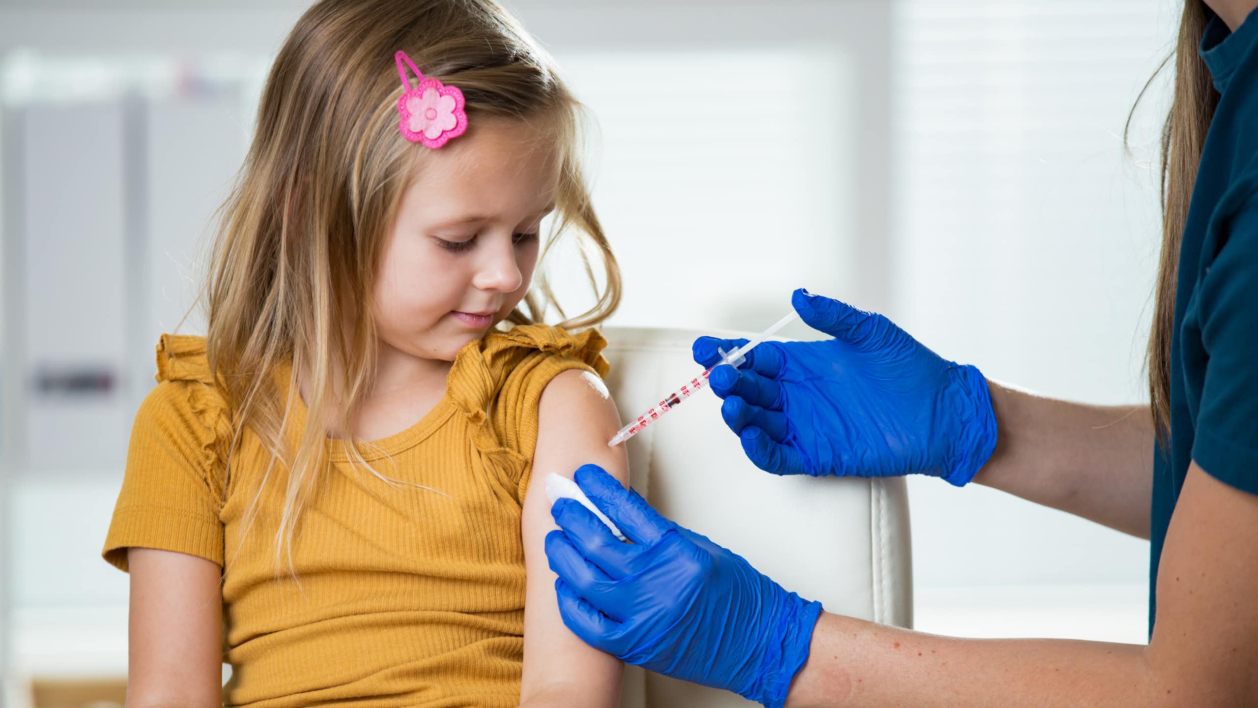 Nurse wearing surgical gloves gives a shot to a school-age girl who sits in a chair.