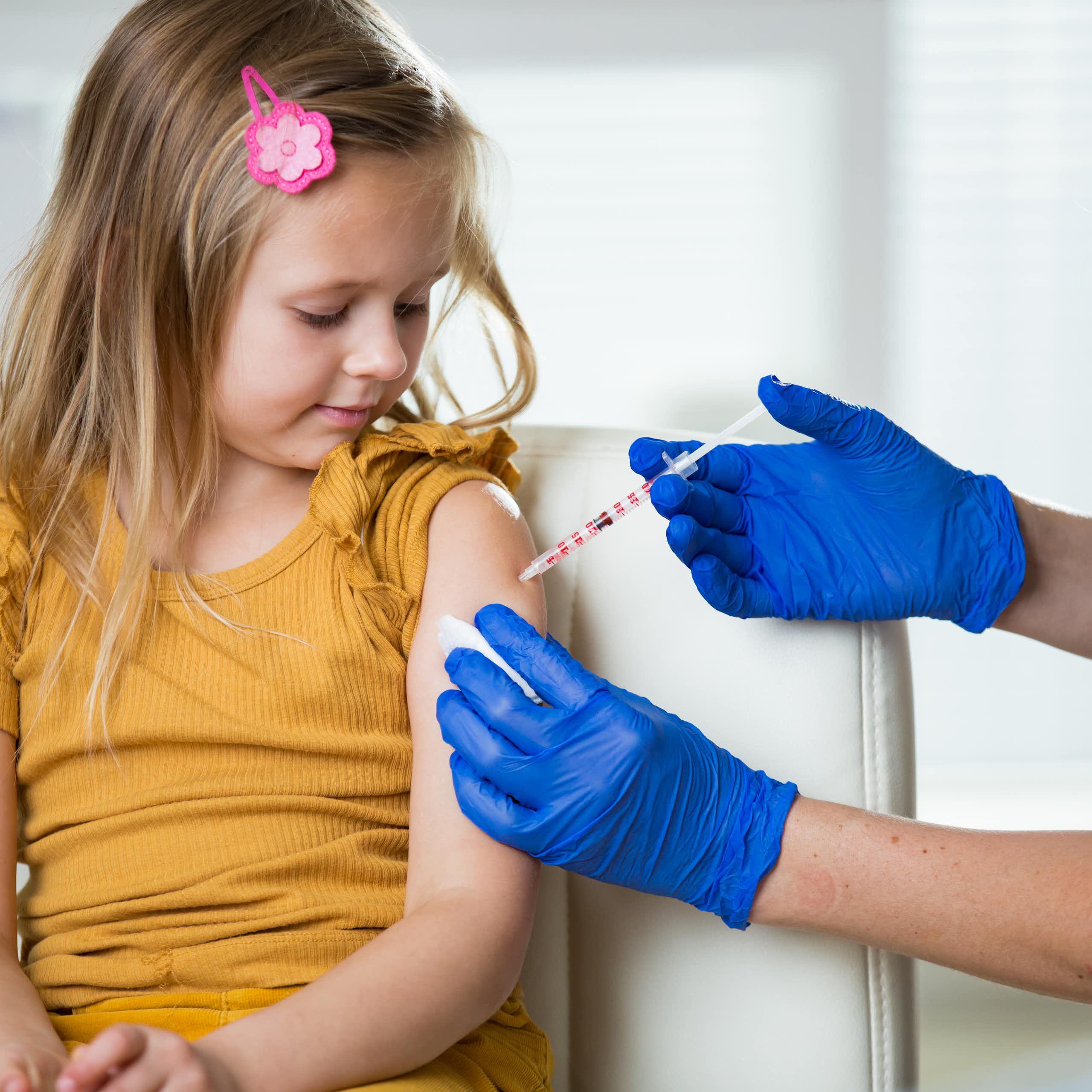 Nurse wearing surgical gloves gives a shot to a school-age girl who sits in a chair.