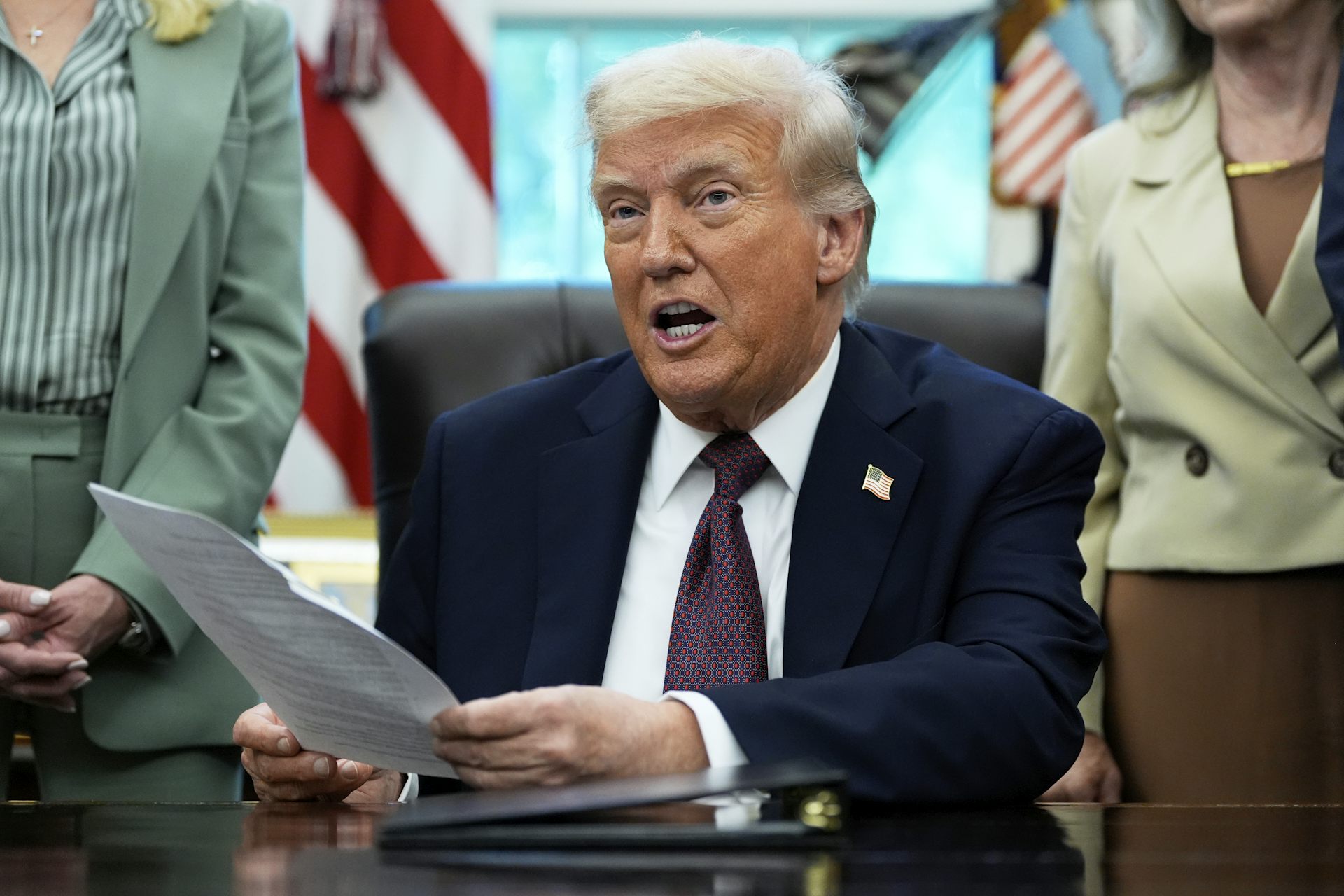 An older white man with white hair in a navy suit speaks while seated at a desk