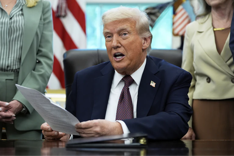 An older white man with white hair in a navy suit speaks while seated at a desk