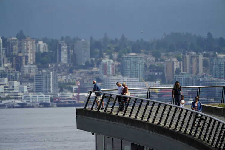 people walk along a waterfront, buildings in the background on the opposing shore