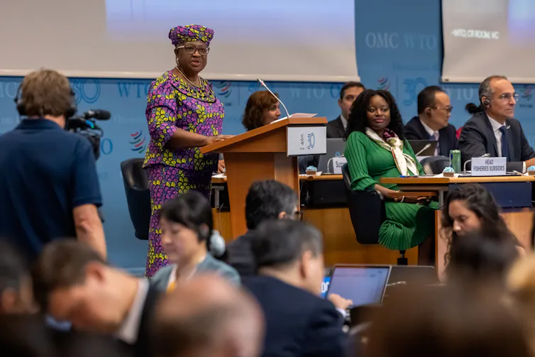 A black woman in a purple and yellow nigerian outfit speaks at a podium, other people are seated behind her and in the audience