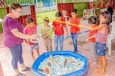 Foto mostra crianças em volta de uma piscina de plástico, pescando insetos ao lado de uma professora