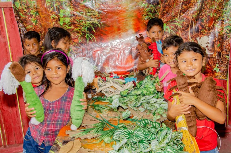 Foto mostra várias crianças em volta de uma mesa com materiais que representam um igarapé amazônico. Elas seguram insetos de pelúcia