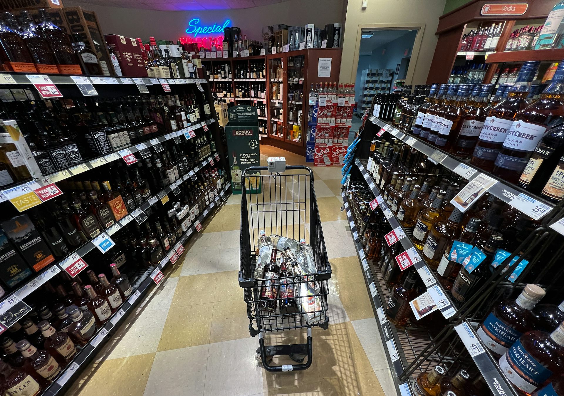 A shopping cart with several bottles in it, in the aisle of a liquor store