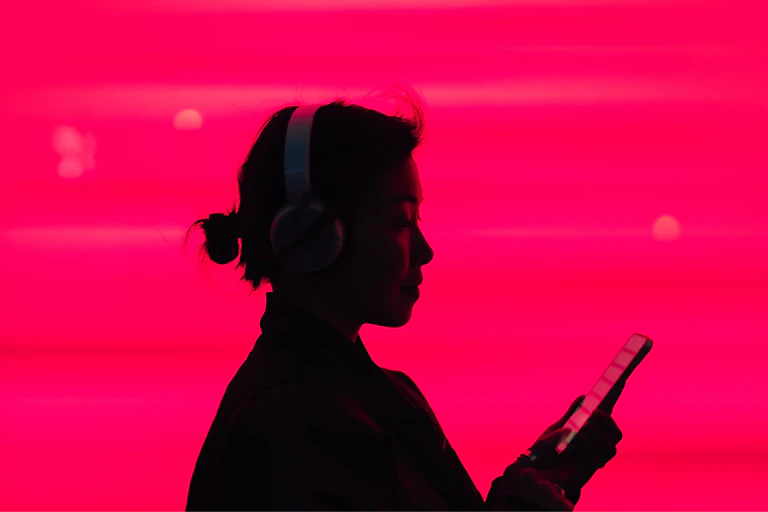 Silhouette of a person wearing headphones and looking at a phone, against a pink background