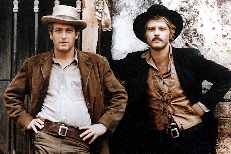 Two men dressed as smart cowboys posing cockily outside a stable.